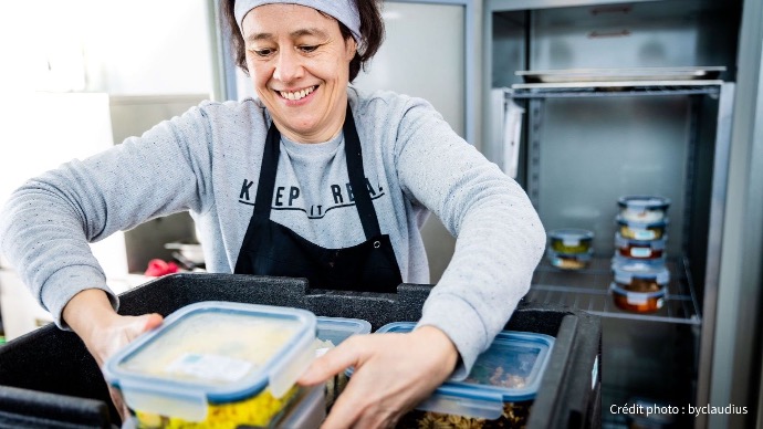 Une femme souriante, portant un tablier noir, organise des boîtes de repas préparés dans un grand bac noir, dans un environnement de cuisine professionnelle.